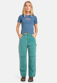 T-shirt bleu court avec logo brodé, associé à un pantalon cargo teal taille haute et à jambes larges, présentant des détails de poches et des coutures visibles.