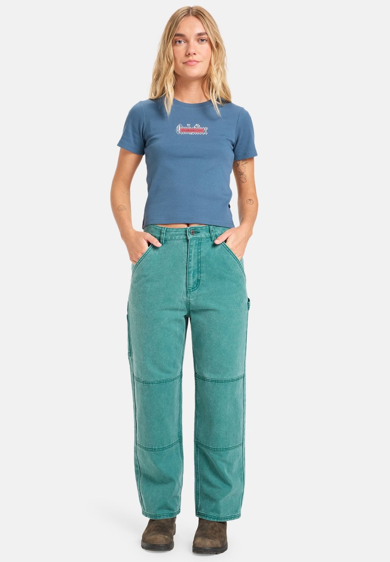 T-shirt bleu court avec logo brodé, associé à un pantalon cargo teal taille haute et à jambes larges, présentant des détails de poches et des coutures visibles.