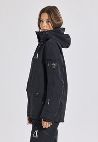 Giacca nera impermeabile con cappuccio, tasche con zip, polsini regolabili e dettagli con logo. Presenta una vestibilità comoda e un tessuto resistente.