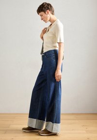 Woman with short hair wearing a cream short-sleeve ribbed top, wide-leg dark blue jeans with rolled cuffs, brown shoes, and silver hoop earrings.