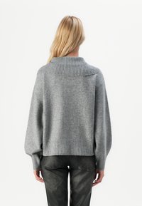 En blond kvinne som vender bort fra kameraet, har på seg en løs, grå ribbestrikket genser med bred krage og svarte teksturerte bukser mot en hvit bakgrunn.