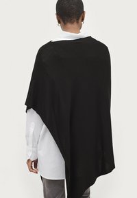Sort poncho-lignende overtøj lavet af let materiale, med en asymmetrisk kant og båret over en hvid skjorte med krave.