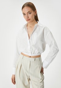 Camicia bianca corta con bottoni frontali, colletto e taschino singolo. Presenta una vita elastica e maniche lunghe. Abbinata a pantaloni leggeri a righe.