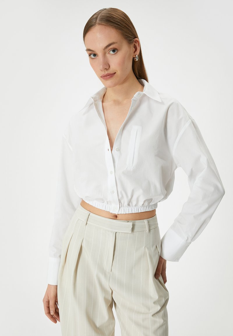 Camicia bianca corta con bottoni frontali, colletto e taschino singolo. Presenta una vita elastica e maniche lunghe. Abbinata a pantaloni leggeri a righe.