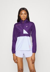 Chaqueta ligera en color púrpura y azul claro con un diseño de media cremallera, puños elásticos y un logo visible. Presenta acentos de bloques de color angulares.