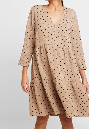 Beige V-neck dress with black polka dots, long sleeves, and tiered skirt worn by a person against a plain background.