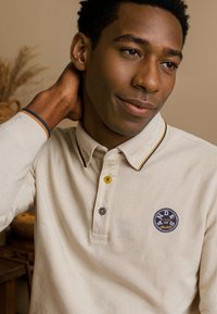 Beige poloshirt met lange mouwen en een kraag, voorzien van een marineblauw en mosterdgeel gestreept accent. Knopdetails en logo patch op de borst.