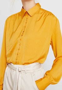 Žlutá saténová halenka s knoflíkovou légou, řasenými rameny a dlouhými rukávy. Kombinována s krémovými plisovanými kalhotami a zavazovacím pasem.