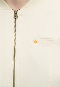 Κοντινή λήψη ενός κρεμ μπουφάν με μπρούντζινο φερμουάρ και κεντημένο λογότυπο "Schott" με ένα μικρό πορτοκαλί αστέρι στο στήθος.