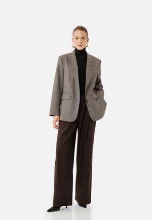 Grau karierter Blazer mit Reverskragen, zwei Fronttaschen und lockerer Passform. Kombiniert mit hochgeschnittenen braunen Hosen und schwarzen spitzen Schuhen.