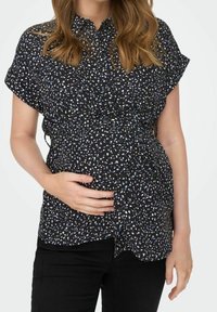 Blouse noire boutonnée avec manches courtes, présentant un motif tacheté blanc et bleu clair, conçue pour une taille cintrée, portée avec un pantalon noir.