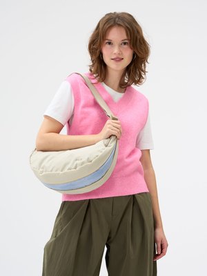 Jeune femme portant un pull sans manche rose sur un t-shirt blanc, un pantalon vert olive, tenant un sac bandoulière beige et bleu clair en forme de croissant.
