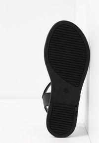 Semelle de sandale noire avec motifs de prise texturés appuyée verticalement contre un mur d'angle blanc et un sol.