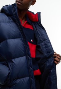 Persona che indossa una giacca a piumino blu navy sopra una maglietta a righe blu navy e rosse con un piccolo logo rettangolare sul petto.