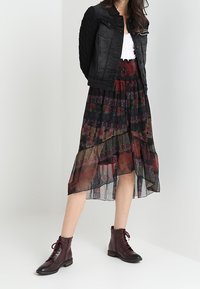 Nainen, jolla on päällään musta farkkutakki, valkoinen toppi, kukkakuosisesta läpinäkyvästä mustasta midi-hameesta sekä tummanpunaiset nauhaliimatut nilkkurit.