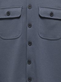 Σκούρο μπλε ύφασμα με διαγώνια υφή, με μια σειρά από μαύρα κουμπιά στο κέντρο και δύο τσέπες με πατούρα και κουμπί κοντά στο επάνω μέρος.