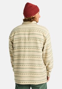 Chemise à longues manches, à motifs crème avec des designs géométriques verts. Devant à boutons, col et texture douce. Bonnet en tricot rouge porté.