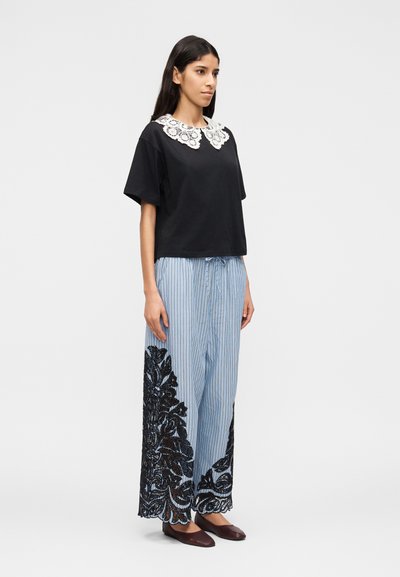 Femme debout portant un haut noir à manches courtes avec un col en dentelle blanche et un pantalon large à rayures bleues avec des détails en dentelle noire florale.