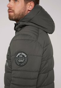 Chaqueta de plumas gris con capucha, que presenta paneles acolchados y un parche redondo con logo en la manga izquierda. El material parece suave y duradero.