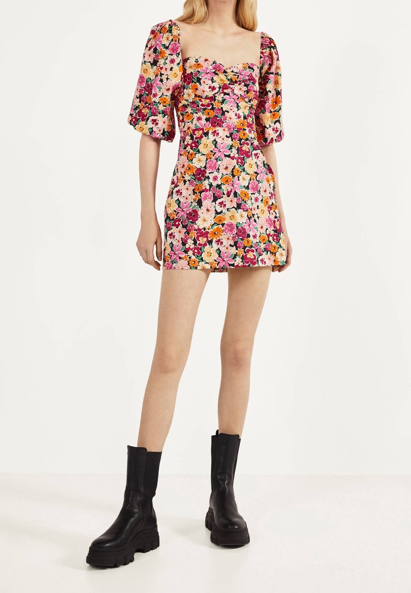 Femme portant une robe courte et ajustée à motifs floraux avec des manches bouffantes et des bottes noires épaisses, sur un fond uni.
