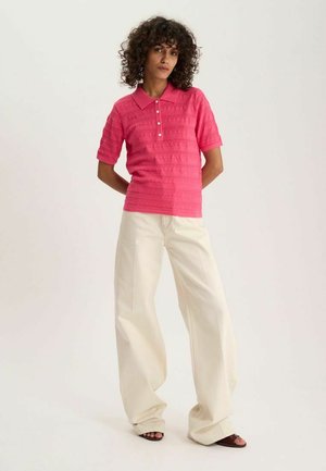 Femme aux cheveux bouclés debout portant un polo rose texturé à manches courtes et un pantalon large crème avec des chaussures à bout ouvert.