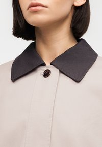 Femme portant un manteau beige avec un grand col marron foncé et un bouton rond en écaille fixé au niveau du cou.