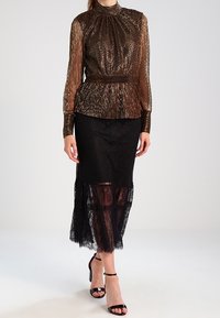 Blusa lunga a maniche corte color bronzo metallizzato abbinata a una gonna midi in pizzo nero con una sezione inferiore trasparente e sandali con cinturino alla caviglia.
