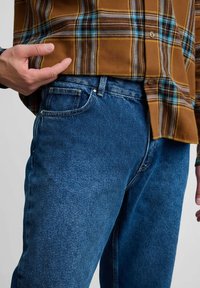 Brązowa koszula w kratę z turkusowymi i żółtymi akcentami, noszona na niebieskich dżinsach z denimu. Dżinsy mają klasyczny krój i widoczne detale przeszyć.