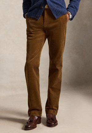 Homme portant un pantalon en velours côtelé marron, un pull bleu foncé, des mocassins en cuir marron à pampilles, et une montre-bracelet, les mains dans les poches.