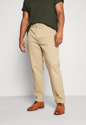 Mężczyzna ubrany w beżowe spodnie chinos, ciemnozieloną koszulkę z podwiniętymi rękawami i brązowe skórzane eleganckie buty, stojący na jednolitym tle.