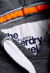 Tela de sudadera gris con capucha, que presenta una etiqueta interior blanca con texto en azul oscuro y un acento naranja de Superdry en el cuello.
