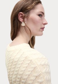 Maglione lavorato a maglia crema con un motivo a treccia; modello che indossa orecchini lisci di forma astratta in bianco sporco; vista di profilo.