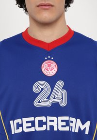 Blauw shirt met rode kraag, met een witte "24" ontwerp, "ICECREAM" tekst en een rond roze patch met een lachend gezicht en sterren.