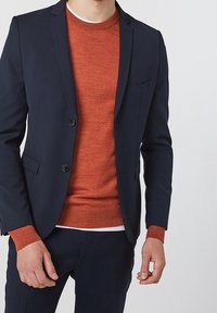 Marineblå kavaj med två knappar och bröstficka, lagd över en orange crewneck-tröja, i kombination med mörka byxor. Slät tygstruktur.