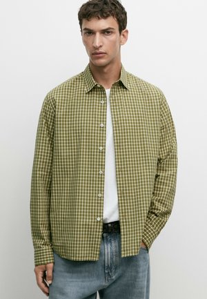 Giovane uomo che indossa una camicia a quadri verde e bianca sbottonata sopra una t-shirt bianca, con jeans azzurri e un'espressione facciale neutra.