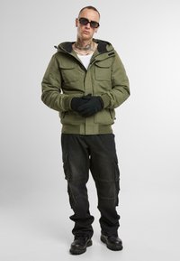 Chaqueta verde oliva con múltiples bolsillos, forro negro, puños acanalados, combinada con pantalones cargo negros y botas negras. Los guantes son de punto negro.