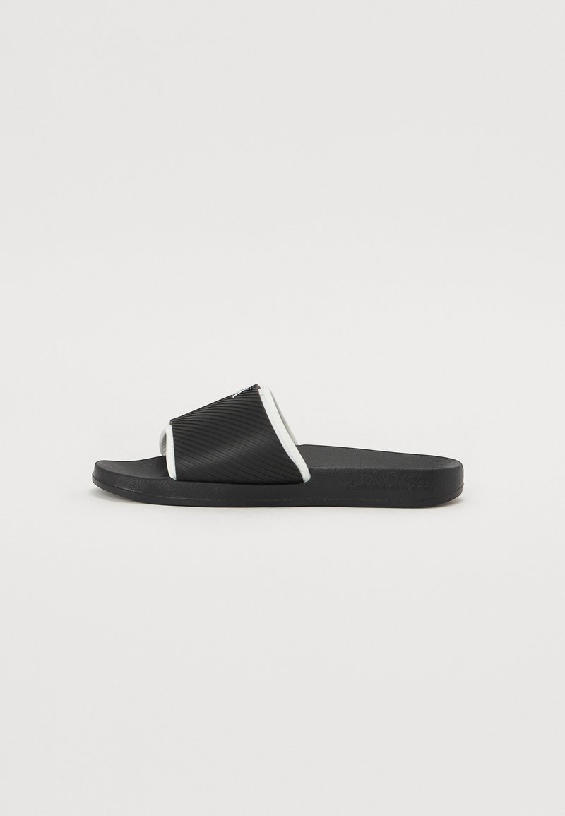 Black slide sandal featuring a ribbed upper with white piping and a textured footbed. Designed for comfort with a flat sole.