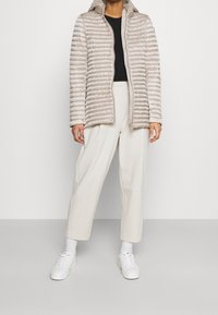 Jasnobeżowa kurtka puchowa z kapturem, z poziomym pikowanym wzorem. Noszona na czarnej bluzce, w połączeniu z kremowymi spodniami o szerokich nogawkach i białymi trampkami.