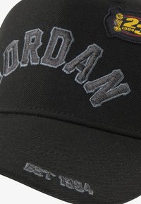 Svart cap laget av bomull med brodert "JORDAN"-tekst i grått, med en fargerik lapp med en basketballtrofé og "23"-merke.