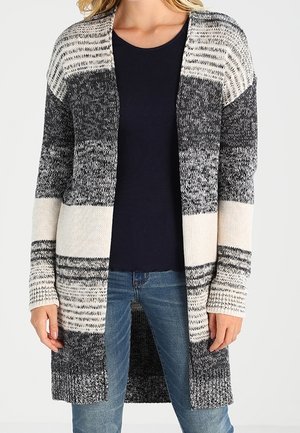 Kobieta ubrana w długi, rozpinany sweter z poziomymi paskami w kolorach czarnym, białym i szarym, na granatowej koszuli i niebieskich dżinsach.