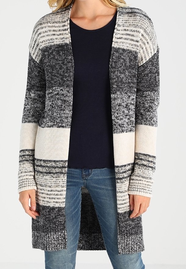 Mujer vistiendo un cárdigan largo de punto abierto al frente con rayas horizontales en negro, blanco y gris sobre una camisa azul marino y vaqueros azules.