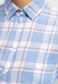 Modrá a bílá kostkovaná košile s červenými akcenty, bavlněný materiál, zapínání na knoflíky a kapsa na hrudi. Měkká textura a neformální vzor.