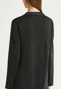 Donna che indossa una maglia nera glitterata a maniche lunghe con colletto, vista da dietro, con capelli castani lunghi e un orecchino d'oro visibile.