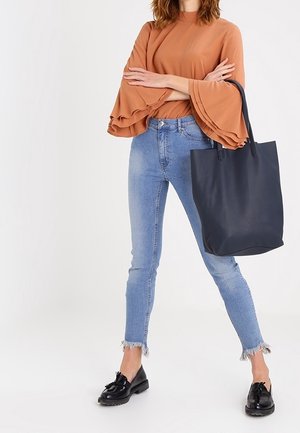 Frau trägt ein orangefarbenes Oberteil mit Rüschen und Glockenärmeln, hellblaue Skinny-Jeans mit ausgefranstem Saum, schwarze Loafers, hält eine große schwarze Tragetasche.