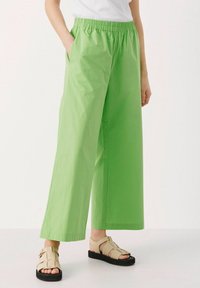 Pantalons larges, légers et verts avec une taille élastique et des poches latérales. Stylisés avec des sandales beiges sur un fond neutre.