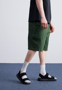 Sandales noires avec des straps réglables, associées à des chaussettes blanches, un short vert et un t-shirt noir, montrées sur une personne en mouvement.
