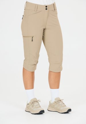 Pantalon beige jusqu'au genou avec une poche zippée sur le côté, porté avec des chaussettes blanches et des chaussures de randonnée beiges, sur une personne debout.