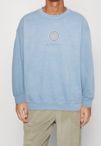 Ljusblå sweatshirt med rund halsringning och ribbad muddar. Har en central grafisk design av en mandala och texten "NATURAL ELEMENTS" nedanför.