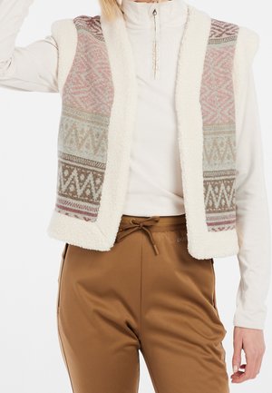 Vrouw draagt wit shirt met lange mouwen, mouwloos fleecevest met patroon in pasteltinten en bruine broek met trekkoord, staat met één arm omhoog.