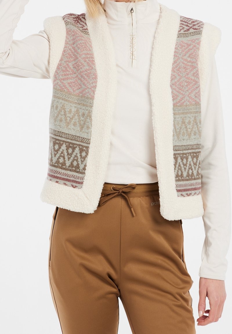 Femme portant un haut blanc à manches longues, un gilet sans manches en polaire à motifs dans des tons pastel et un pantalon marron avec cordon, debout avec un bras levé.